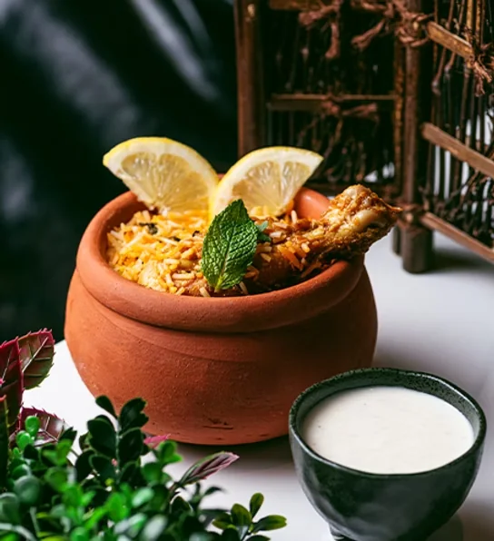 Biryani served in a clay pot with lemon wedges and raita